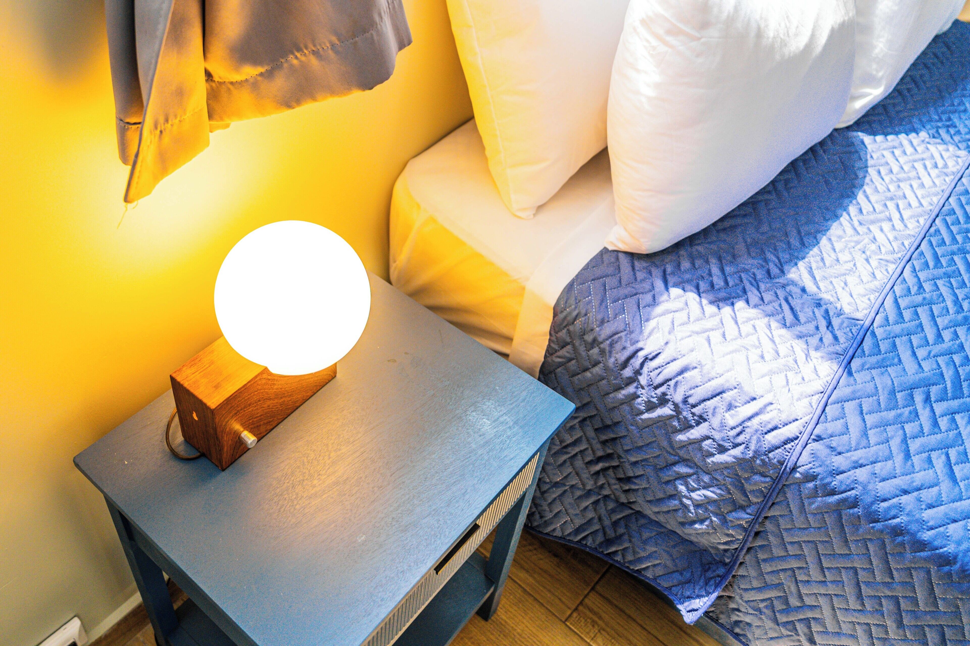 Un lit recouvert d’une couverture matelassée bleue, une table de chevet avec une lampe en bois et un luminaire fixé au mur.