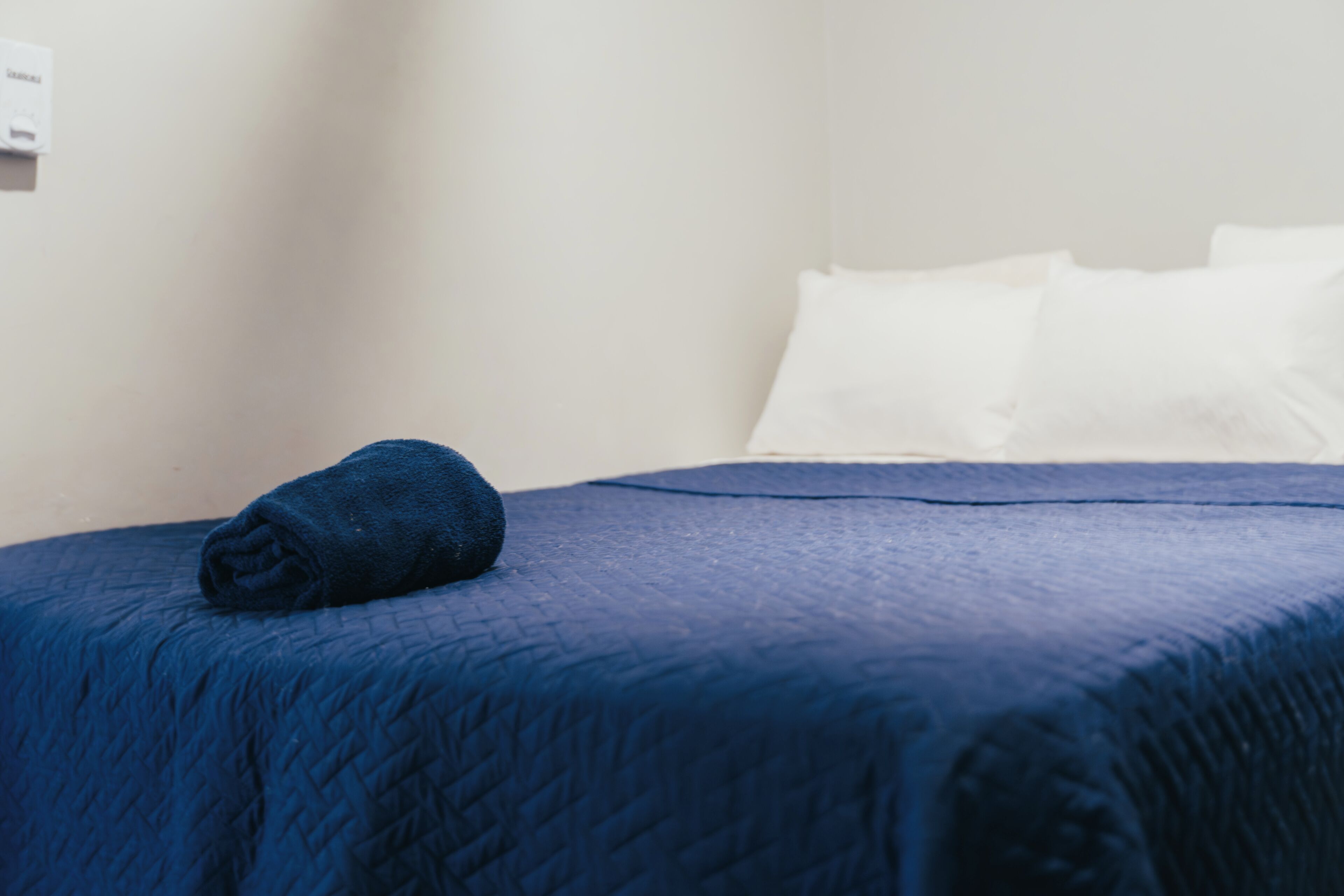 Una cama bien hecha con colcha azul, almohadas blancas y un enchufe de pared.