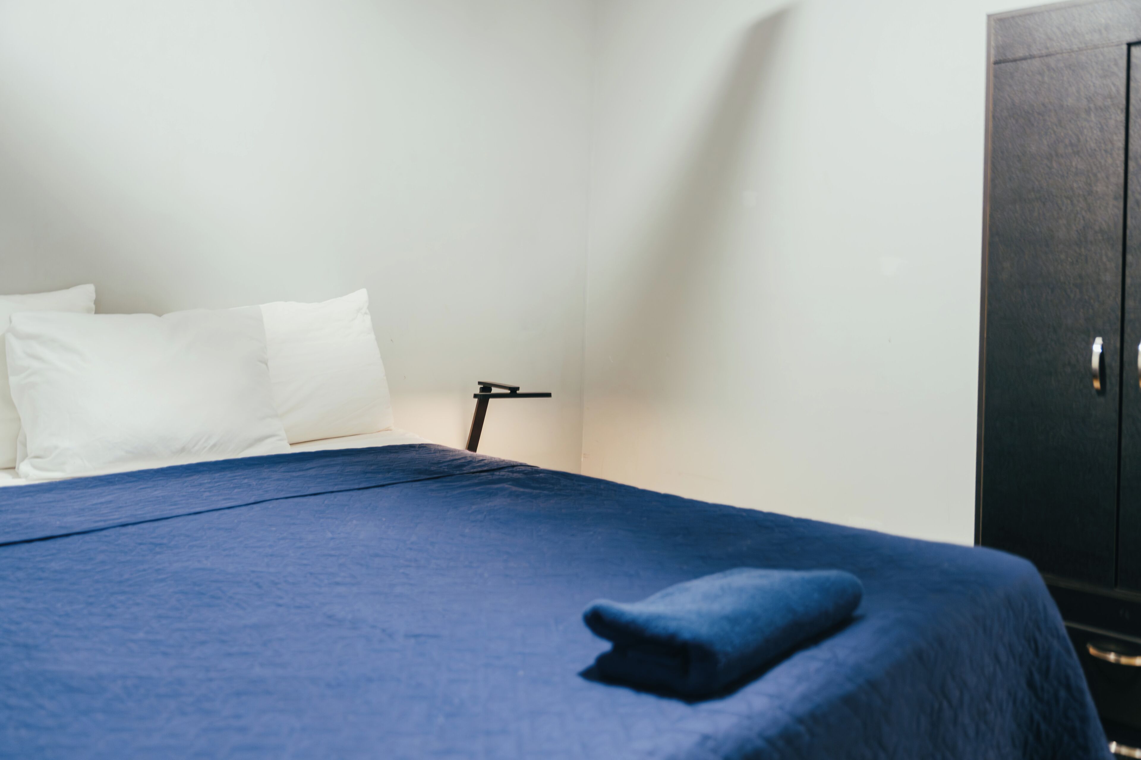 A bed with a blue bedspread, white pillows, and a black nightstand with a lamp.