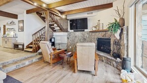 A spacious living area with a stone fireplace, wooden beams, a staircase, and a TV mounted on the wall. - Stylishly Remodeled Mountain Retreat on Eagle Vail Golf Course (Avon)