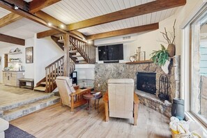Ein geräumiger Wohnbereich mit Kamin aus Stein, Holzbalken, einer Treppe und einem an der Wand befestigten Fernseher.