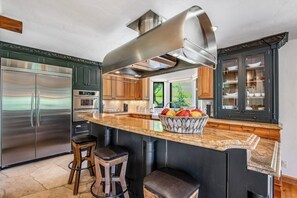 Eine Küche mit einem Edelstahl-Kühlschrank, einem eingebauten Backofen, einer großen Kochinsel mit Obstschale und Holzbarhocker.