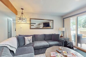 A modern living room with a grey sectional sofa, a round coffee table, a TV, and a large window. - Lovely condo with fireplace, mountain view, full kitchen, & skiing nearby (Avon)