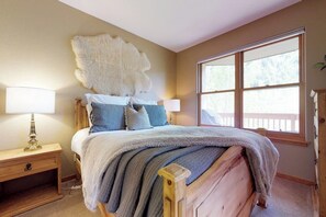 Ein Schlafzimmer mit großem Fenster, einem Bett mit vielen Kissen, einem Nachttisch aus Holz und einem Fellteppich an der Wand.
