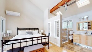 A bedroom with a bed, bedside table, bench, and a bathroom with a sink and mirror. - Townhome with stunning mountain views, fireplace, deck, enclosed patio, & W/D (Avon)