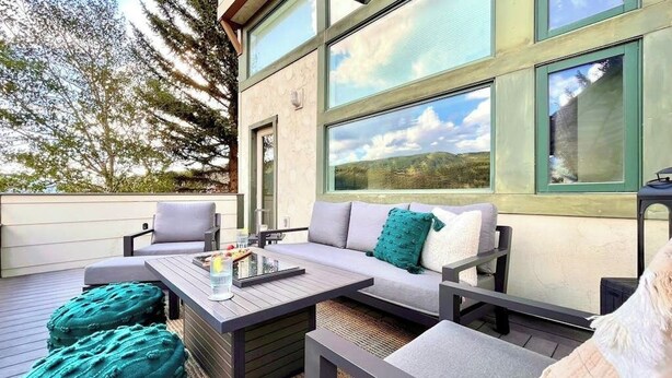 An outdoor seating area with a sofa, chairs, and ottomans, a table with a tray, and large windows offering a view of the landscape. - Townhome with stunning mountain views, fireplace, deck, enclosed patio, & W/D (Avon)