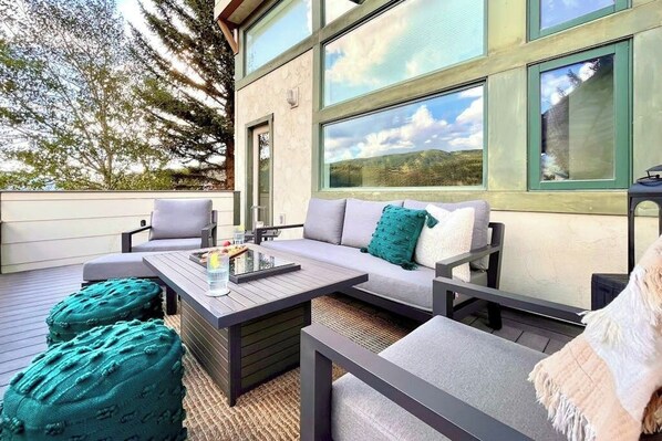 Ein Außenbereich mit einer Couch, Sesseln, Poufs, einem Tisch mit Tablett und großen Fenstern mit Blick auf die Landschaft.