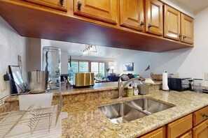 A kitchen with granite countertops, stainless steel sink, wooden cabinets, and a modern light fixture. - Mountain view condo with large balcony, pool & hot tub - steps to ski shuttle (Vail)