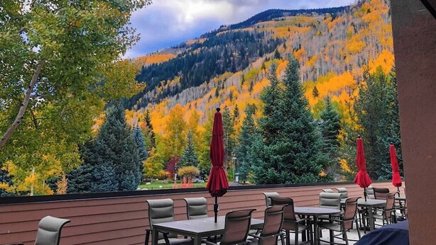 An outdoor patio with tables and chairs, red umbrellas, and a view of a forested mountain. - Mountain view condo with large balcony, pool & hot tub - steps to ski shuttle (Vail)