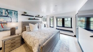 Un dormitorio con cama, mesitas de noche, estante en la pared y un amplio ventanal.