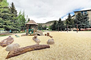 Ein Spielplatz mit einer Holzspielanlage, Sandboden und umliegenden Bäumen.
