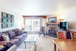 Ein Wohnzimmer mit einer Couch, Sesseln, einem Couchtisch, einem Kamin, einem Fernseher und einem Balkon.