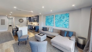 Ein modernes Wohnzimmer mit einer Couch, Sesseln und einem Couchtisch. Im Hintergrund ist eine Küche mit Schränken und einer Mikrowelle zu sehen.