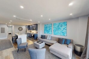 Ein modernes Wohnzimmer mit einer Couch, Sesseln und einem Couchtisch. Im Hintergrund ist eine Küche mit Schränken und einer Mikrowelle zu sehen.