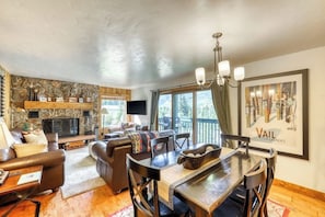 A cozy living room with a fireplace, leather sofas, a dining table, and a large window offering a view of the outdoors. - Upscale condo on shuttle route to Vail w/ a full kitchen, shared pool & tennis (East Vail)