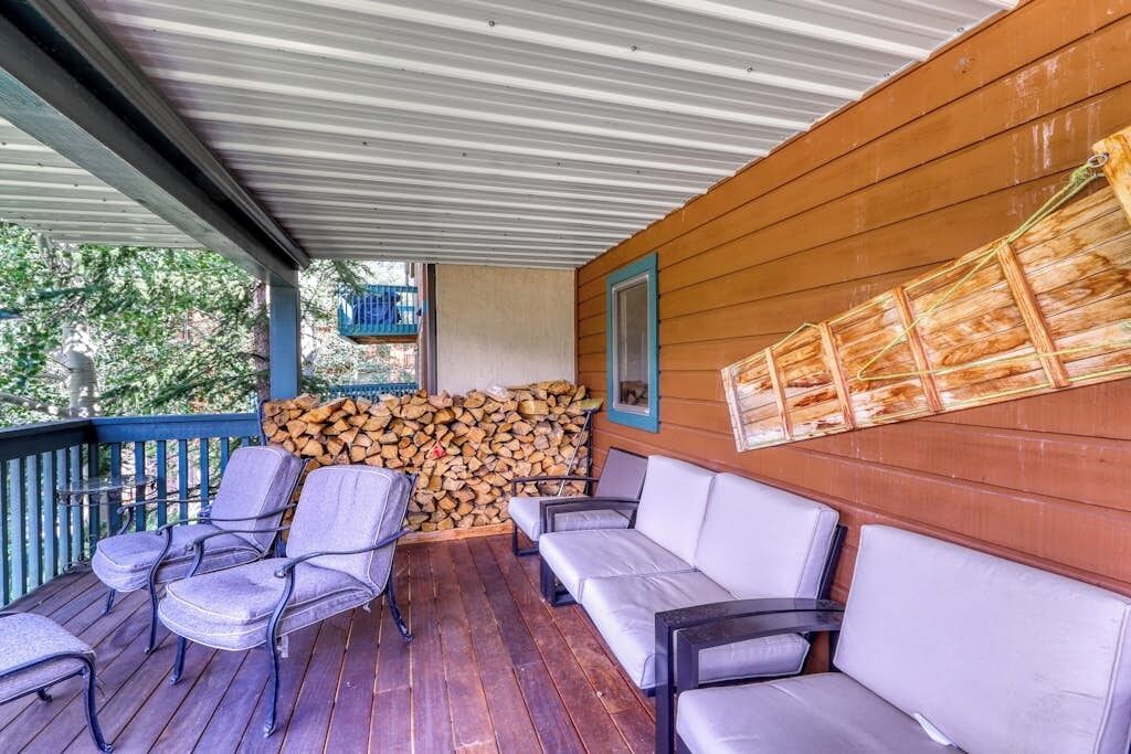 A covered porch with wooden walls, a stack of firewood, and two rows of chairs and a sofa.
