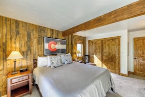 A bedroom with a large bed, wooden walls, a ceiling with exposed beams, a nightstand with a lamp, and a painting above the bed. - Upscale condo on shuttle route to Vail w/ a full kitchen, shared pool & tennis (East Vail)