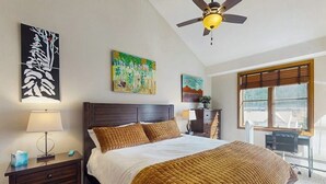 Un dormitorio con cama, mesita de noche, lámpara, ventilador de techo, obra de arte, ventana y escritorio.