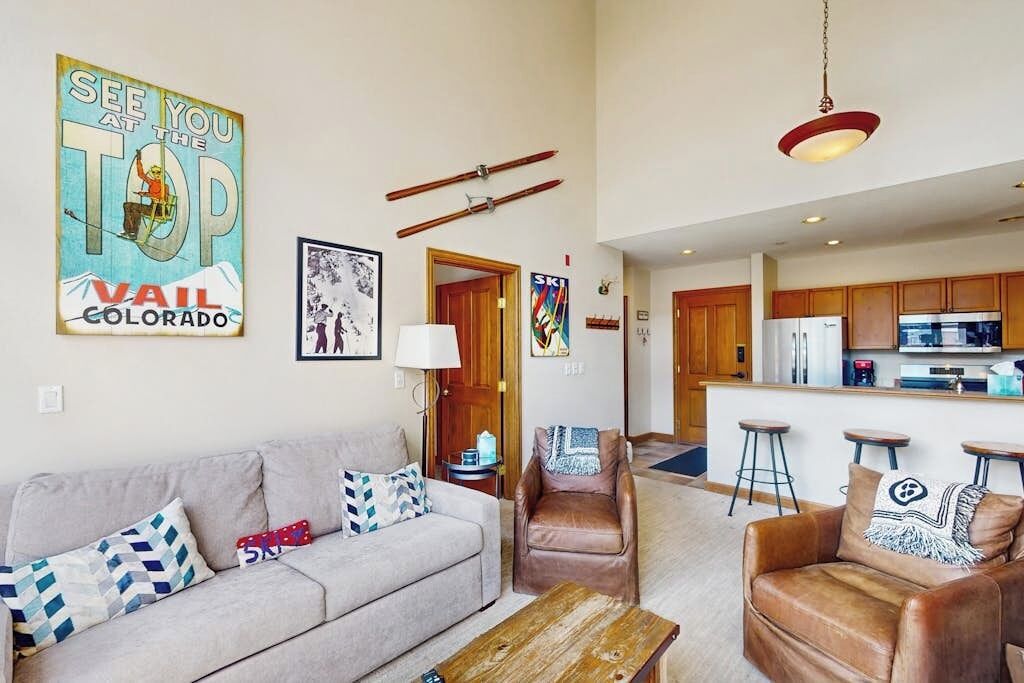 Ein Wohnzimmer mit einer Couch, einem Couchtisch und einem braunen Ledersessel. Im Hintergrund ist eine Küche mit einem Kühlschrank und einer Bar zu sehen.