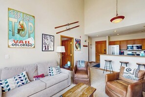 A living room with a sofa, a coffee table, and a brown leather chair. There is a kitchen in the background with a refrigerator and a bar counter. - Conveniently located condo with kitchen, fast wifi & balcony & mountain views (Avon)