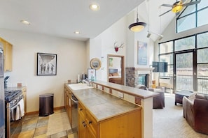 A modern kitchen with a central island, a dining area with a fireplace, and large windows offering a view. - Conveniently located condo with kitchen, fast wifi & balcony & mountain views (Avon)