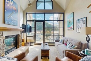 A spacious living room with a fireplace, large windows, a sofa, and a coffee table. - Condo with A/C and heated parking, under half a mile to Riverfront Gondola (Avon)