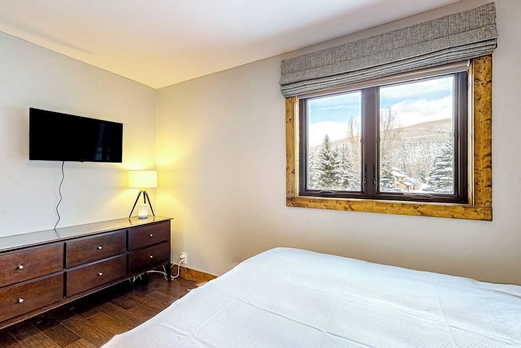 A bedroom with a bed, a dresser, a TV mounted on the wall, and a window with a view of snow-covered trees.