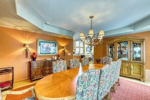 A dining room with a large wooden table, floral upholstered chairs, a chandelier, a cabinet with glass doors, and a framed picture on the wall.