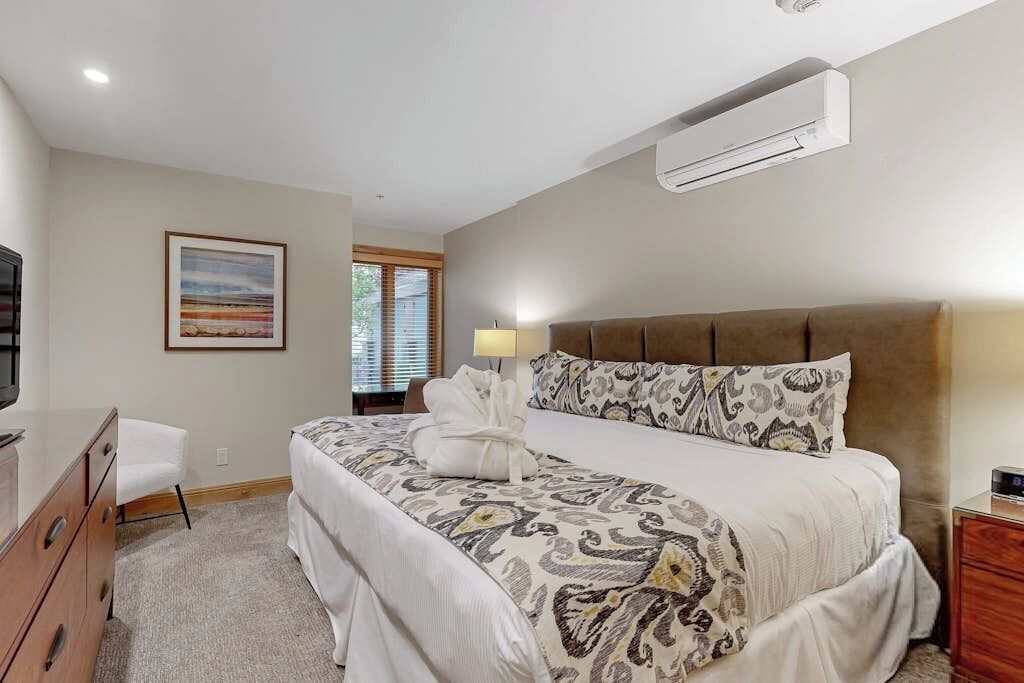 A bedroom with a bed, a bedside table, a lamp, a dresser, a window with blinds, and an air conditioning unit.