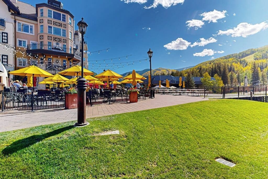 An outdoor café with yellow umbrellas, tables, and chairs, surrounded by buildings and a grassy area.