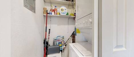 A narrow bathroom with a white wall, a shelf with cleaning supplies, a bucket labeled 'CERAMIC WALL TILE,' and a white washing machine.