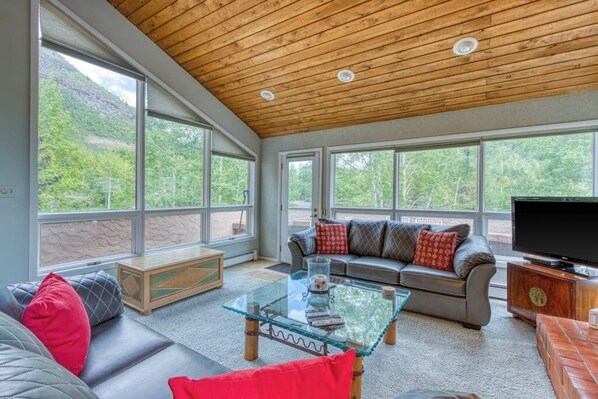 A spacious living room with a large window, a wooden ceiling, a sofa, a glass coffee table, and a television. - Secluded valley home w/ private balcony, grill, shared tennis - close to trails (East Vail)