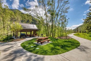 A picnic area with a wooden table and benches, surrounded by a grassy lawn and trees, with a building in the background. - Secluded valley home w/ private balcony, grill, shared tennis - close to trails (East Vail)