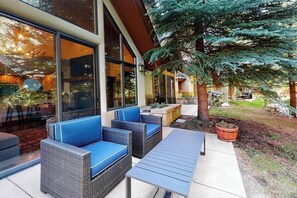 Eine Terrasse mit Korbmöbeln und einem Tisch, ein großer Baum und ein Gebäude mit großen Fenstern.