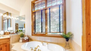 Une salle de bain avec une grande baignoire, des stores en bois aux fenêtres, un miroir avec éclairage intégré et une plante en pot.