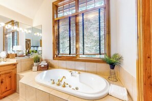 Ein Badezimmer mit einer großen Badewanne, Holzjalousien, einem beleuchteten Spiegel und einem Blumentopf.