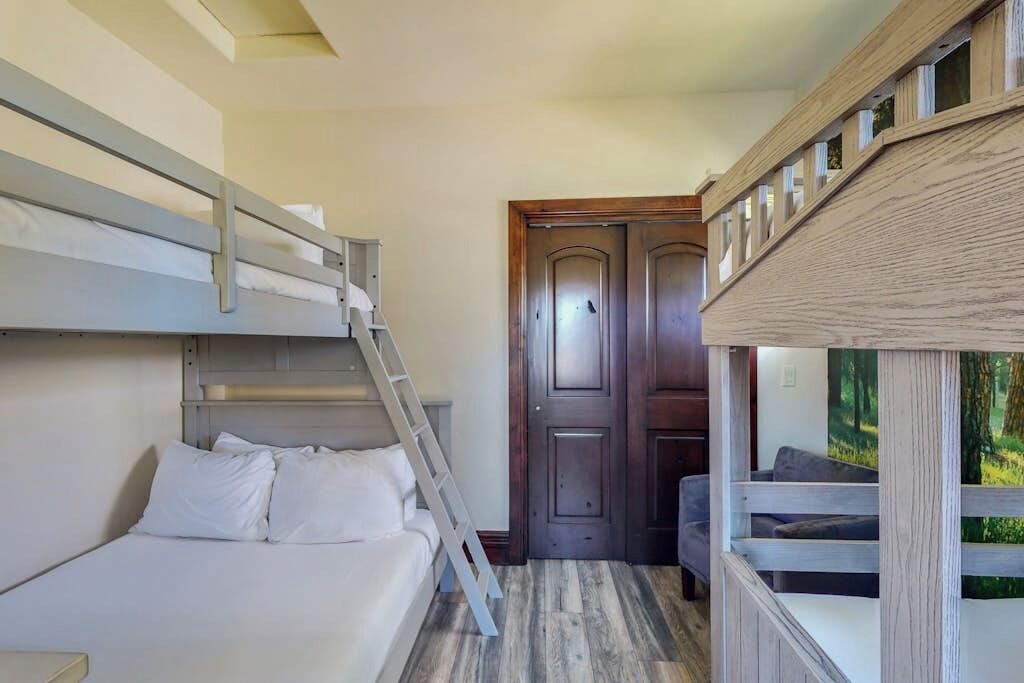 A bunk bed room with a ladder, a wooden door, and a view of greenery.