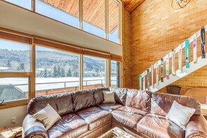 Ein Zimmer mit einem ledernen Ecksofa, großen Fenstern mit Blick auf eine verschneite Landschaft und einer Treppe mit Ski-Ausrüstung.