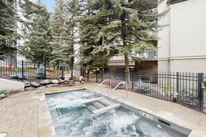 A hot tub with a metal ladder, surrounded by a paved patio, a black metal fence, and tall evergreen trees. - Spacious condo w/ full kitchen, shared hot tub & perfect location (Beaver Creek)