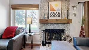 A living room with a stone fireplace, a sofa, a coffee table, and a view of trees outside. - Mountain-view condo with outdoor pool, fireplace, patio, pool table & W/D (Edwards)
