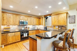 A kitchen with wooden cabinets, a granite countertop, stainless steel appliances, and a bar area with chairs. - Second-floor retreat near the slopes with grill, fireplace, pool, & hot tub (Edwards)