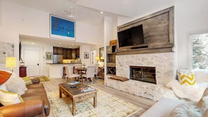 A spacious living area with a fireplace, a television mounted above it, and a kitchen in the background. - All-season condo near ski lifts & golf course, with pool, & hot tub (Edwards)
