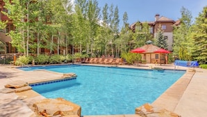 A swimming pool with lounge chairs and a red umbrella, surrounded by trees and buildings. - All-season condo near ski lifts & golf course, with pool, & hot tub (Edwards)
