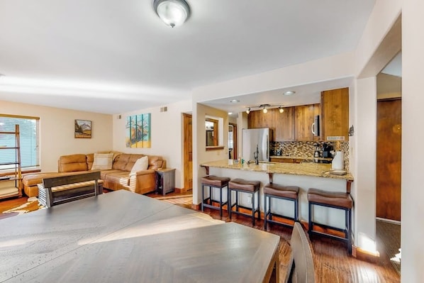 A spacious living area with a large dining table, a kitchen with wooden cabinets, and a bar area with stools. - Riverfront, ground-floor condo with fireplace & fishing access - near Riverwalk (Edwards)