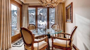 Ein Essbereich mit einem Glastisch und Holzsesseln, einer Chandelierleuchtre und Blick auf eine verschneite Landschaft durch große Fenster.