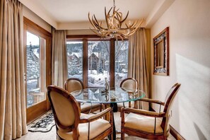 A dining area with a glass table and wooden chairs, a chandelier, and a view of a snowy landscape through large windows. - Winter Condo in Ski-In/Ski-Out Complex w/Fireplace/Free WiFi/Shared Pool/Hot Tub (Vail)
