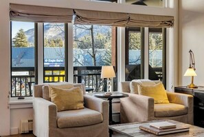 A room with two armchairs, a small table, and a lamp. Large windows offer a view of mountains and a building with signage. - Ski-in/out home with fireplace, AC, high-speed WiFi, shared pool/hot tub/laundry (Vail)