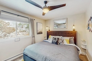 Ein Schlafzimmer mit einem großen Bett, einem Deckenventilator, einem Fenster mit Vorhängen, einem Nachttisch und einem wandmontierten Spiegel.