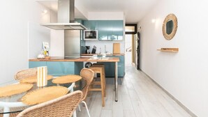 Cocina moderna con una isla central, comedor con sillas de mimbre y un pequeño estante de madera en la pared.