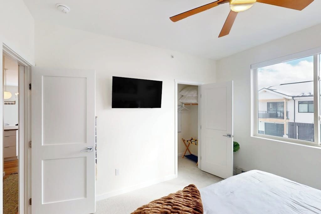A bedroom with a bed, a ceiling fan, a TV, and a window.